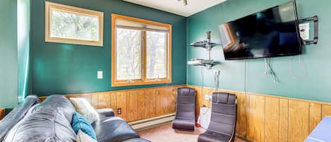A room with a large flat-screen TV mounted on the wall, a wooden paneled wall, two windows, a massage chair, and a massage table.