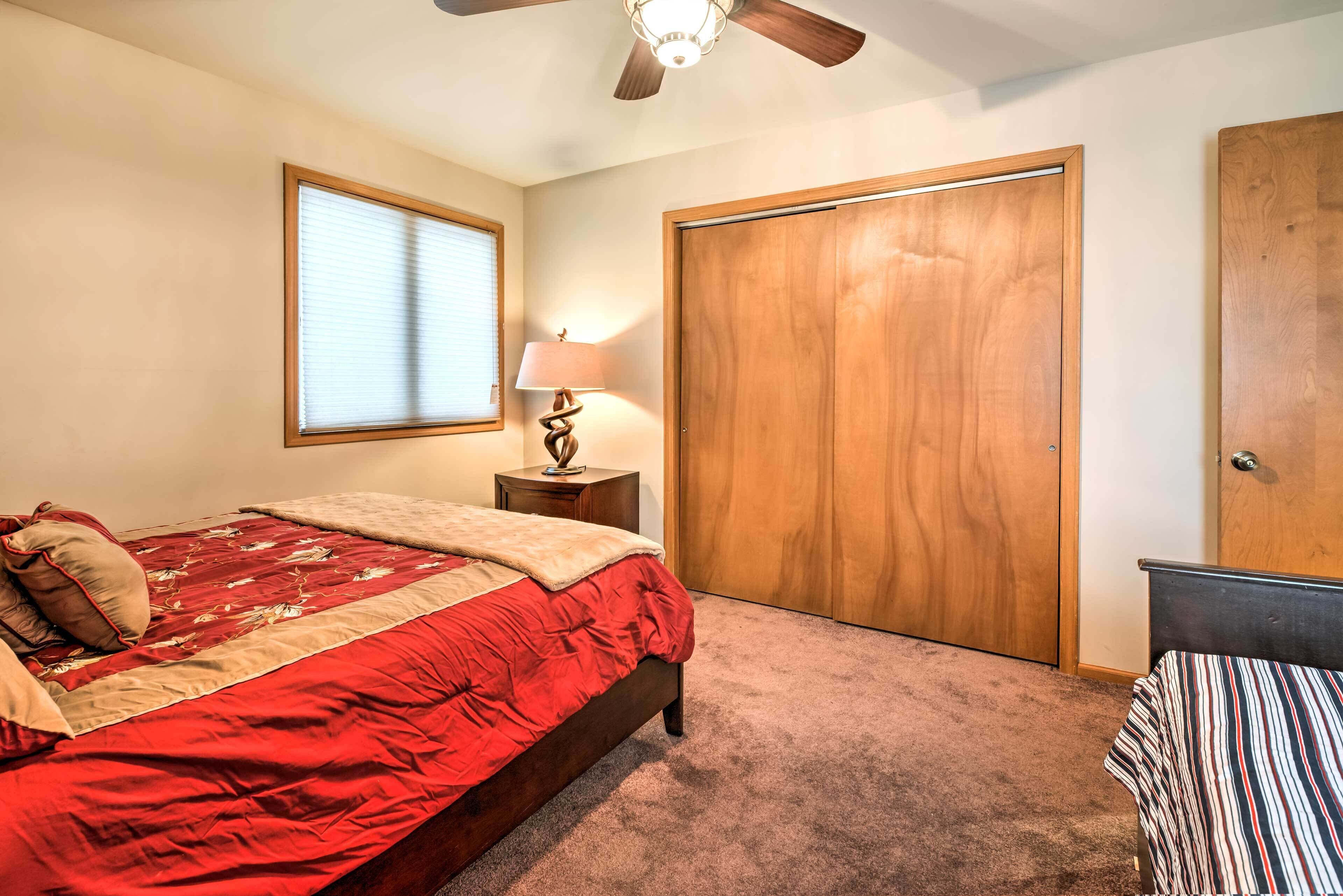 A bedroom with a bed, a nightstand, a lamp, a ceiling fan, a window with blinds, and a wooden wardrobe.