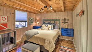 A cozy bedroom with a wooden ceiling, a bed with striped bedding, a nightstand, a crib, and a window with blinds. - Scenic Greers Ferry Cabin w/ Deck + Fire Pit! (Higden)