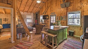 A rustic cabin interior with a wooden ceiling, a stone fireplace, a kitchen area, and a cozy seating area. - Scenic Greers Ferry Cabin w/ Deck + Fire Pit! (Higden)