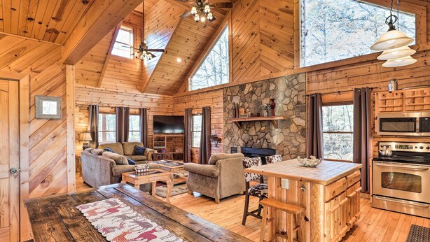 A cozy cabin interior with a stone wall, wooden furniture, a fireplace, and a kitchen area. - Bryson City Cabin w/ Private Hot Tub & Game Room (Bryson City)