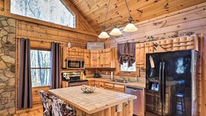 A cabin kitchen with wooden walls, a stone accent wall, a central island, and a black refrigerator. - Bryson City Cabin w/ Private Hot Tub & Game Room (Bryson City)