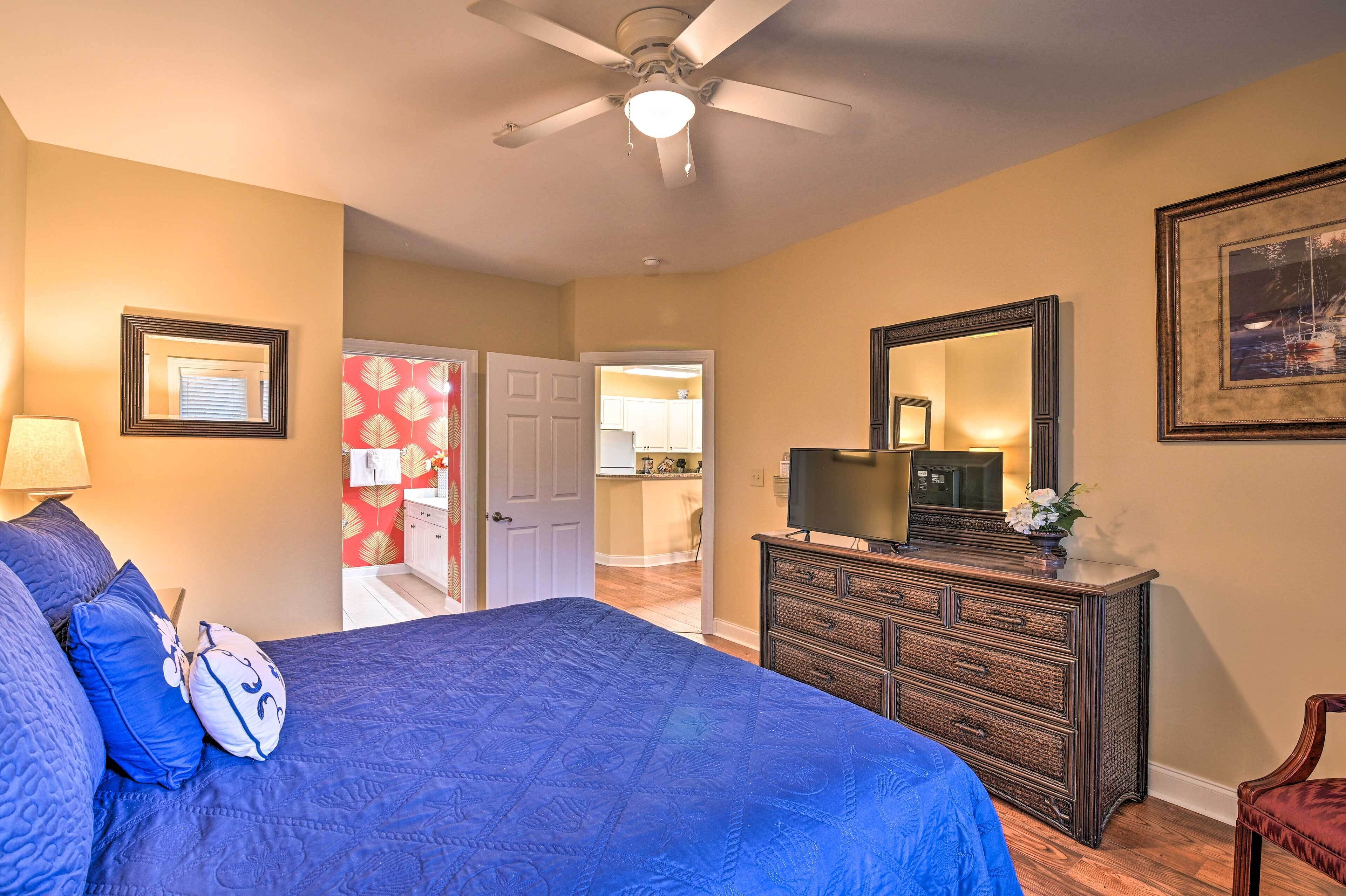 A bedroom with a large bed, a dresser, a ceiling fan, and a door leading to a bathroom.