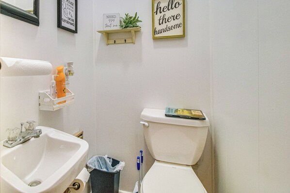 A bathroom with a sink, toilet, and a shelf with toiletries. There are two signs on the wall, one reading "NEED HELP? text me" and another saying "hello there handsome". - Creekfront Yellville Manor w/ Parlors & Fire Pit! (Yellville)