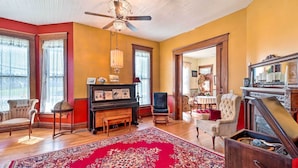 A spacious room with a wooden floor, a large red rug, a piano, and a ceiling fan. - Creekfront Yellville Manor w/ Parlors & Fire Pit! (Yellville)