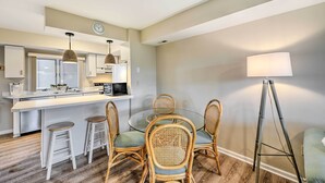 A modern kitchen with a central island, white bar stools, and a dining table with wicker chairs. - Brigantine Condo w/ Beach Access & Pool Views (Brigantine)