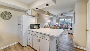 A modern kitchen with a white refrigerator, microwave, and wooden cabinets. There are two pendant lights hanging above the island, and a dining area with a sofa and chairs in the background. - Brigantine Condo w/ Beach Access & Pool Views (Brigantine)