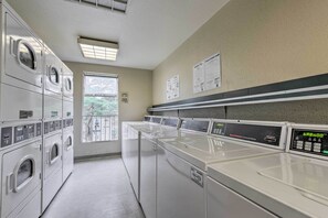 A laundry room with multiple washing machines and dryers, a window with a view of greenery, and informational signs on the wall. - Cali Studio w/ Resort Perks: 6 Mi to San Fran (Daly City)