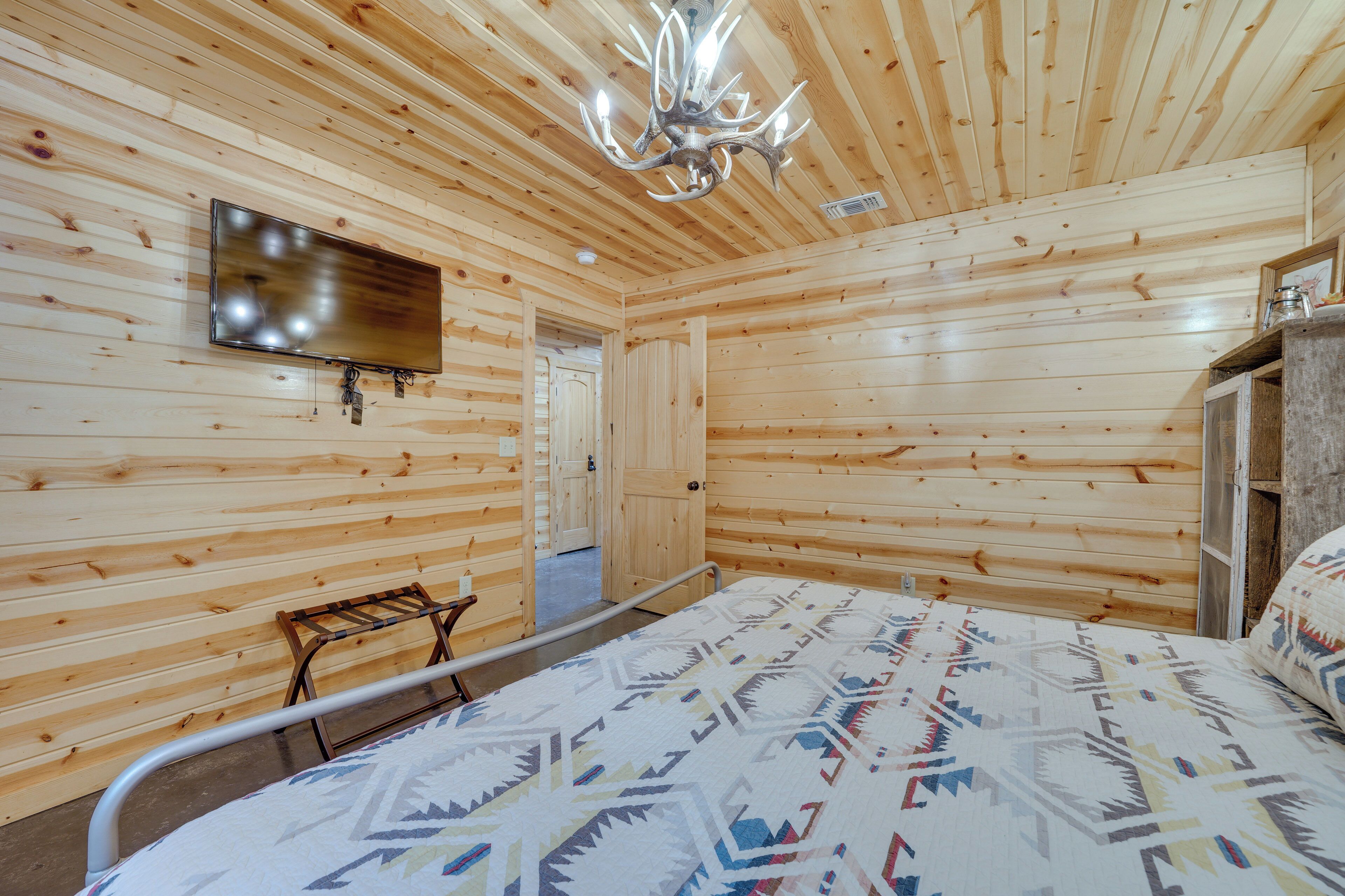 Habitación con paneles de madera, una cama, televisor montado, candelabro y una puerta que da a otra habitación.