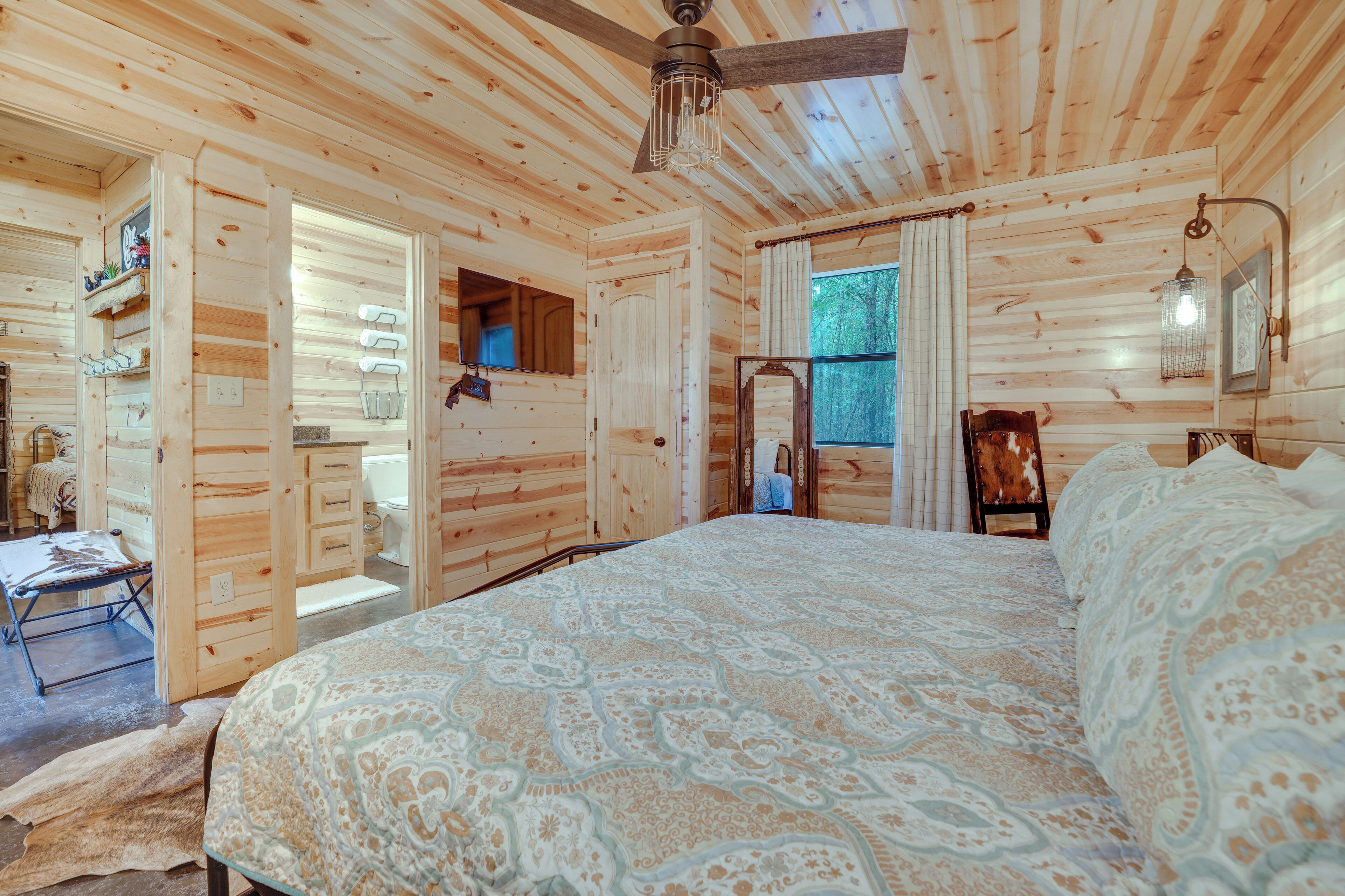 Interior de una cabaña de madera con una cama grande, un ventilador de techo y una ventana con cortinas.