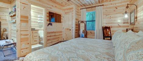 Interior de una cabaña de madera con una cama grande, un ventilador de techo y una ventana con cortinas.