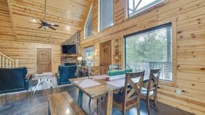 A wooden cabin interior with a dining area, two blue chairs, a wooden table, a ceiling fan, and a stone fireplace. - 'penny Lane Lodge' Broken Bow Cabin < 6 Mi to Lake (Broken Bow)