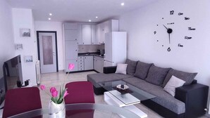 Ein modernes Wohnzimmer mit einem Glastisch, einem grauen Sofa und einem roten Sessel. Im Hintergrund ist eine Küche mit weißen Schränken und einer Mikrowelle zu sehen.