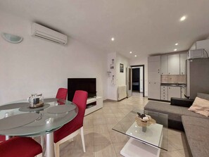 Una sala moderna con una mesa de centro de vidrio, sillas rojas, un televisor de pantalla plana y una zona de cocina con armarios y un refrigerador.