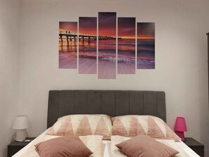 A bedroom with a bed, pillows, a nightstand, a lamp, and a wall-mounted multi-panel sunset painting. - Holiday apartment close to the sea (Malinska)