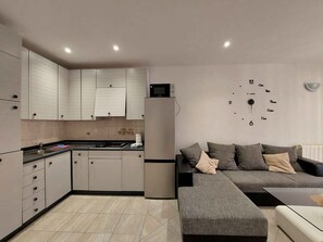 A modern kitchen with white cabinets, a refrigerator, and a microwave. There is a gray sofa with cushions and a small table. - Holiday apartment close to the sea (Malinska)