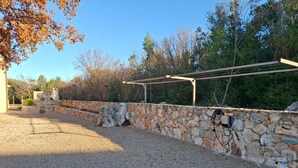 Eine Steinsäule mit einer Pergola, Kiesgrund und Bäumen im Hintergrund.