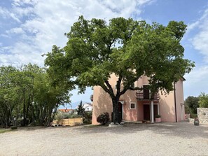 A two-story building with a large tree in front, a gravel driveway, and a clear sky. - Holiday apartment close to the sea (Malinska)