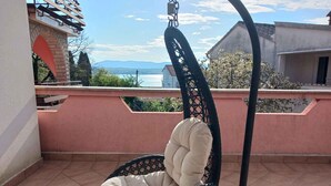 Ein Hängekorbstuhl mit Kissen und einem Buch, platziert auf einem Balkon mit Blick auf ein Gewässer und Berge.
