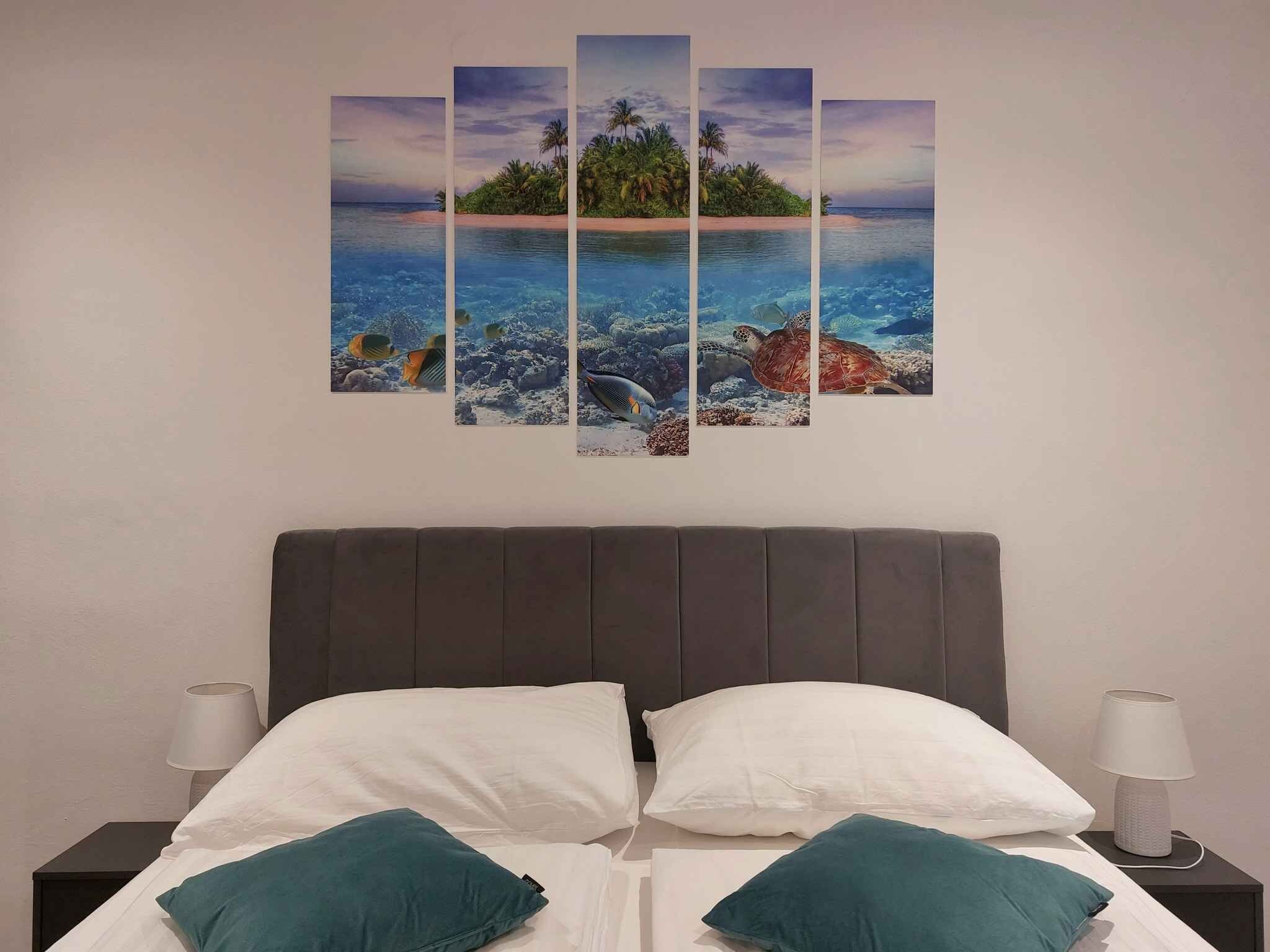 Una cama con almohadas blancas y turquesa, un cabecero oscuro y una obra de arte de varios paneles colgada en la pared que muestra una escena de playa tropical.