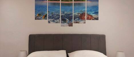 Una cama con almohadas blancas y turquesa, un cabecero oscuro y una obra de arte de varios paneles colgada en la pared que muestra una escena de playa tropical.
