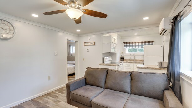 Un salon moderne avec un canapé, un ventilateur de plafond, une horloge et une cuisine en arrière-plan.