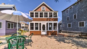 A two-story building with a central entrance, a covered outdoor seating area with chairs and an umbrella, and a sandy ground.