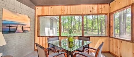 A dining area with a glass table, wooden chairs, and a view of a forest through large windows.