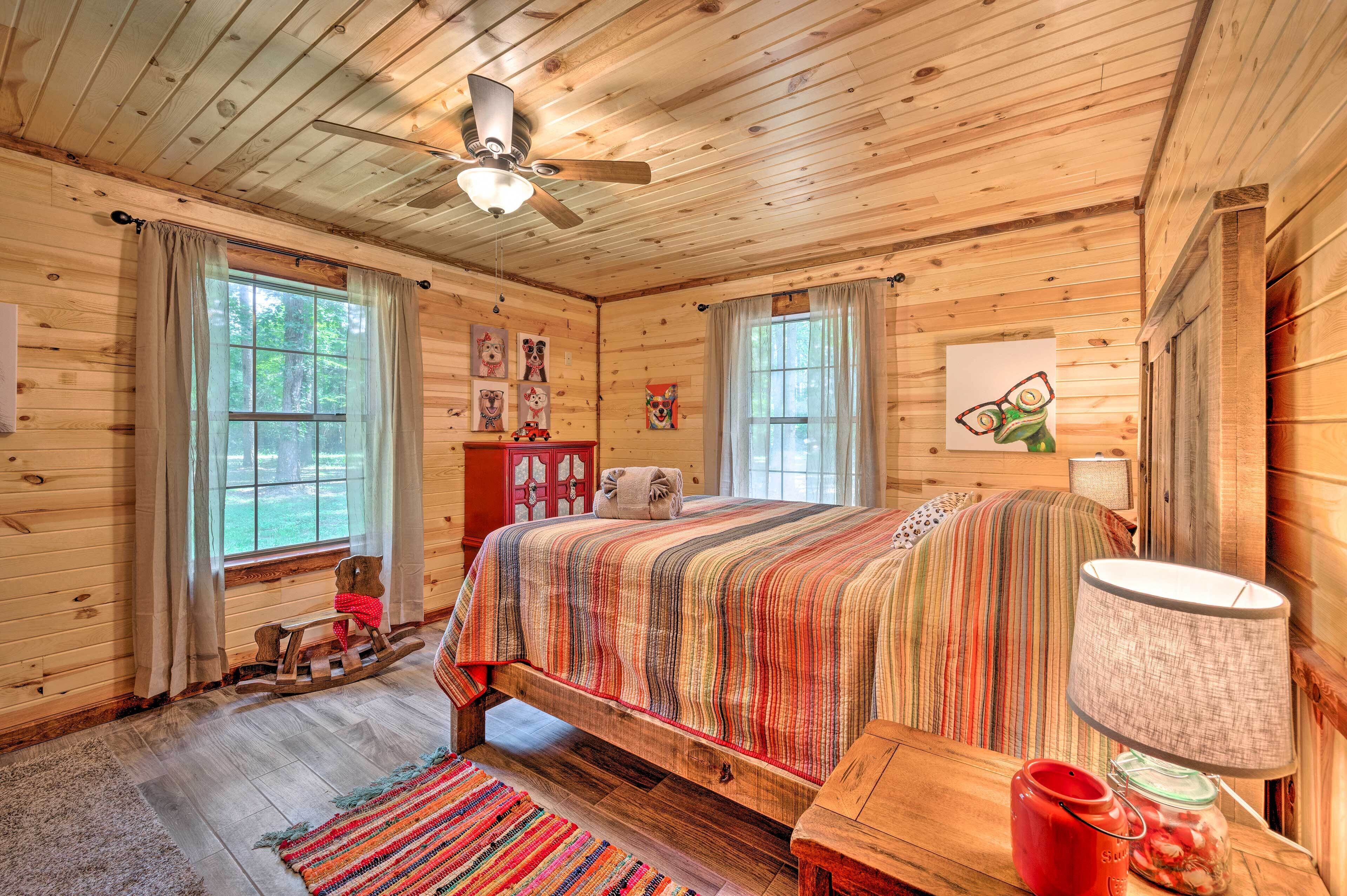 A cozy cabin-style bedroom with a wooden ceiling, a bed with a colorful striped bedspread, a rocking chair, and a small table with a lamp.