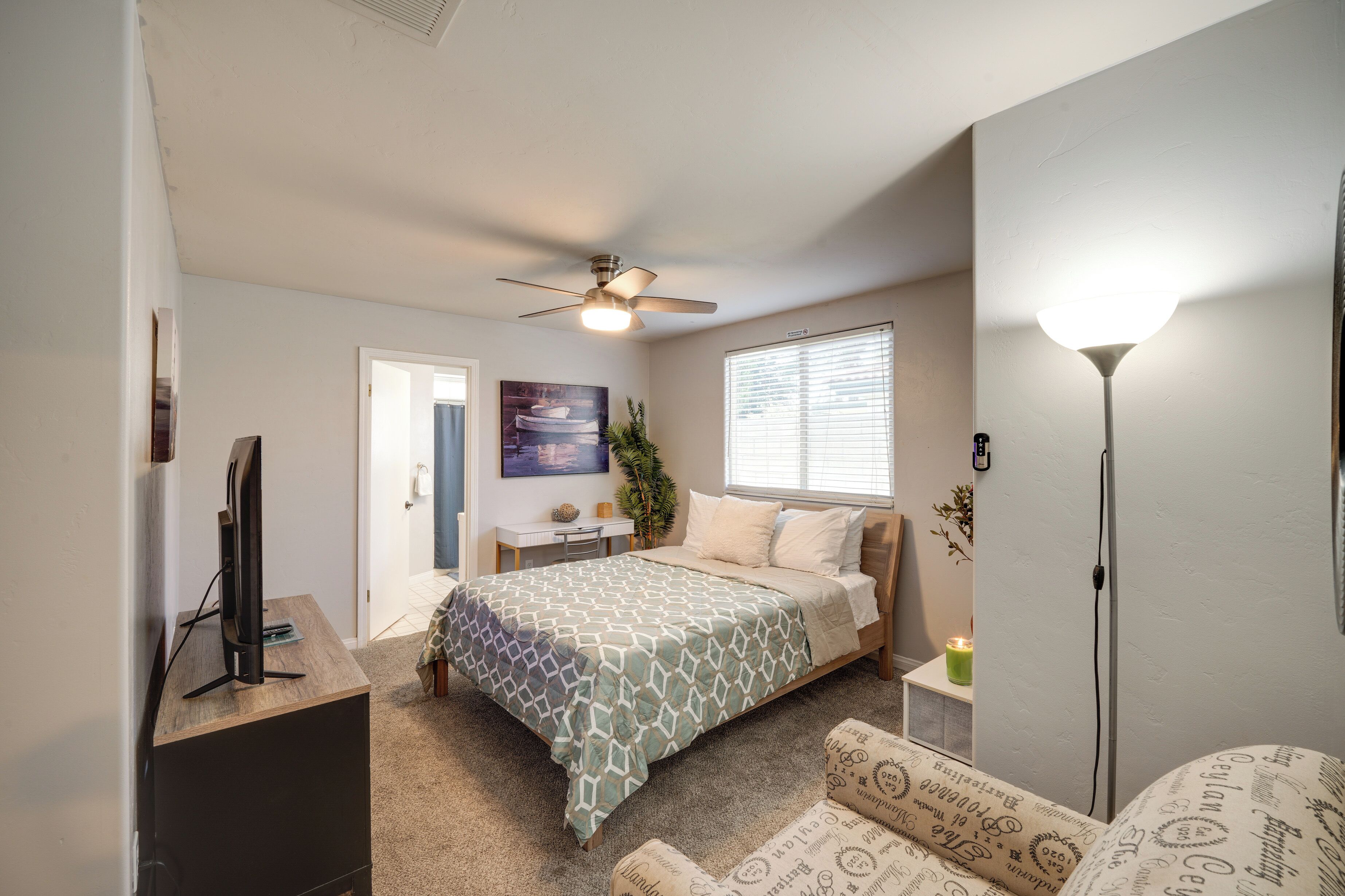Een slaapkamer met een bed, een bureau, een televisie, een plafondventilator, een raam met jaloezieën en een vloerlamp.
