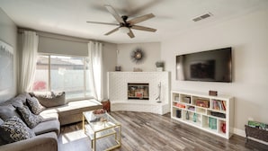 Un salon moderne avec une cheminée en briques blanches, un grand téléviseur à écran plat, un ventilateur de plafond et une bibliothèque.