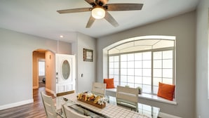 Un coin repas avec une table en verre, des chaises, un ventilateur de plafond et une fenêtre agrémentée de coussins orange.