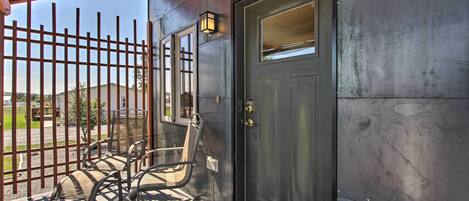 A covered outdoor seating area with metal chairs and a wooden table, a dark metal door with a window, and a metal fence.