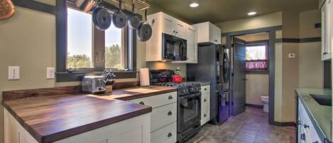 A kitchen with white cabinets, a stainless steel refrigerator, a microwave, a stove, and a hanging pot rack.