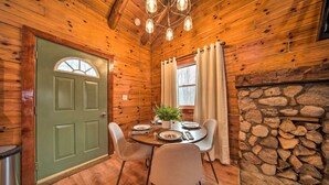 Interior de una cabaña de madera con una mesa de comedor para cuatro, una chimenea de piedra y una puerta verde.