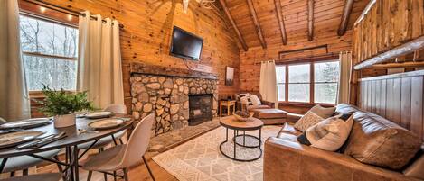 A rustic cabin interior with a stone fireplace, wooden walls, a leather sofa, and a dining area with a table set for four.