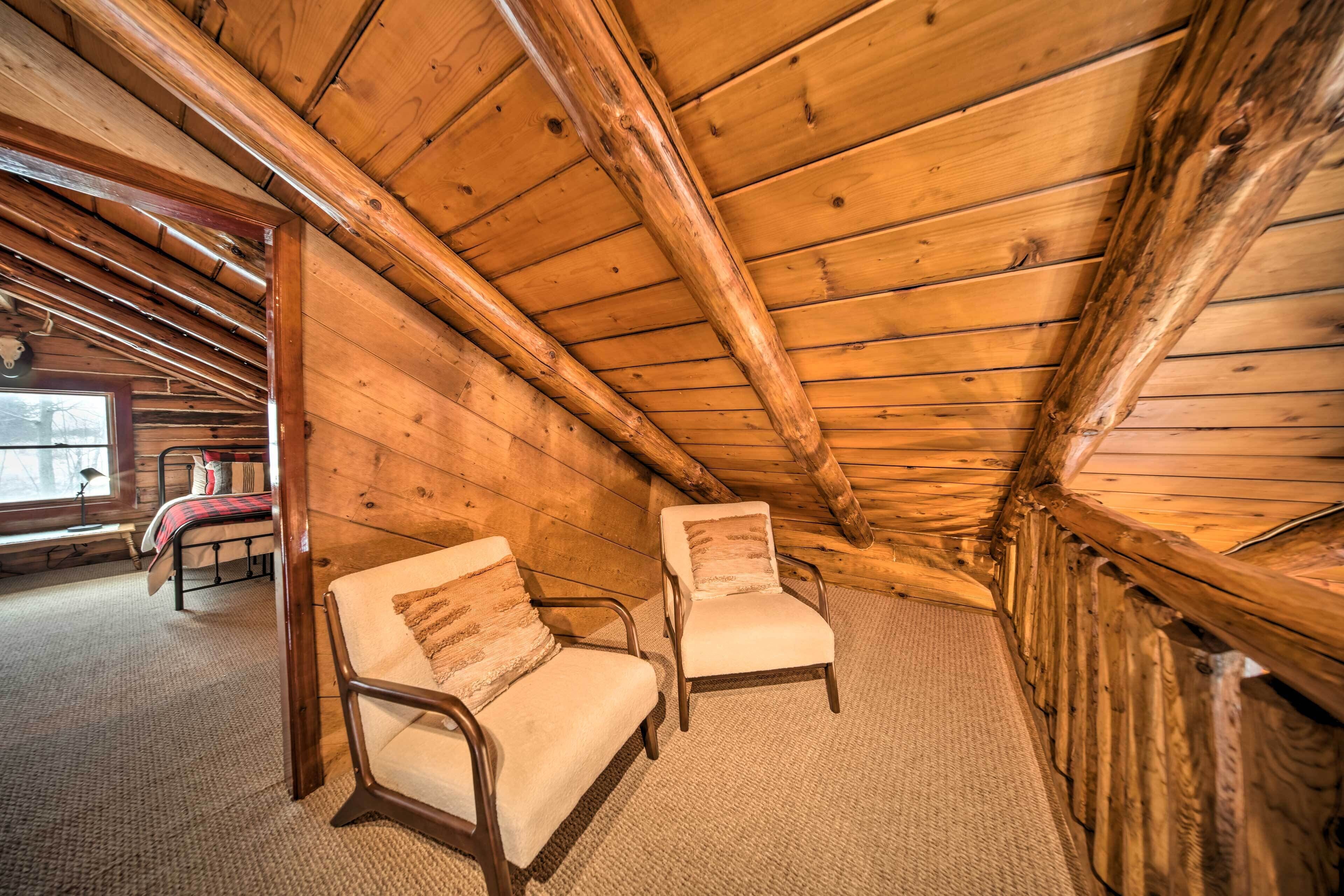 A cozy attic bedroom with wooden walls and a sloped ceiling. There are two upholstered chairs with cushions and a small bed with a red and white checkered blanket.