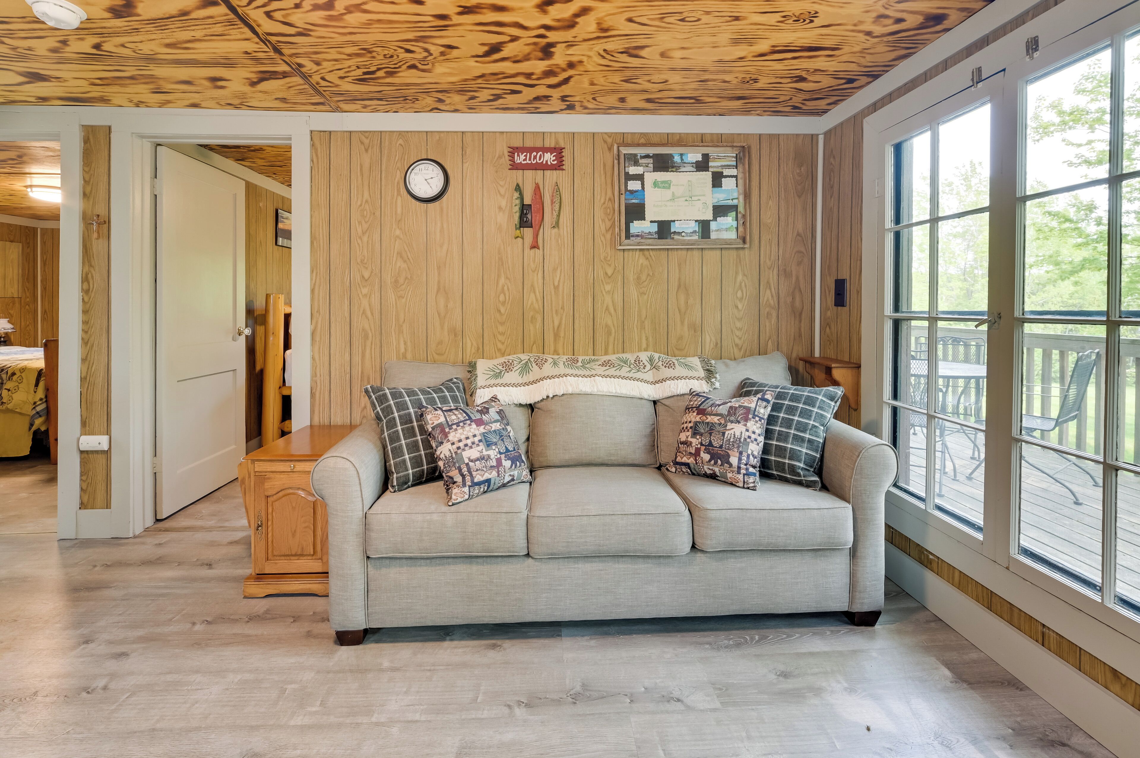 A cozy room with a wooden ceiling, a beige sofa with patterned cushions, a wooden cabinet, and a bulletin board with a welcome sign.