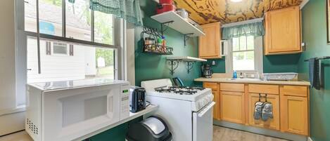A compact kitchen with a white stove, microwave, and sink, wooden cabinets, and a window with a checkered valance.
