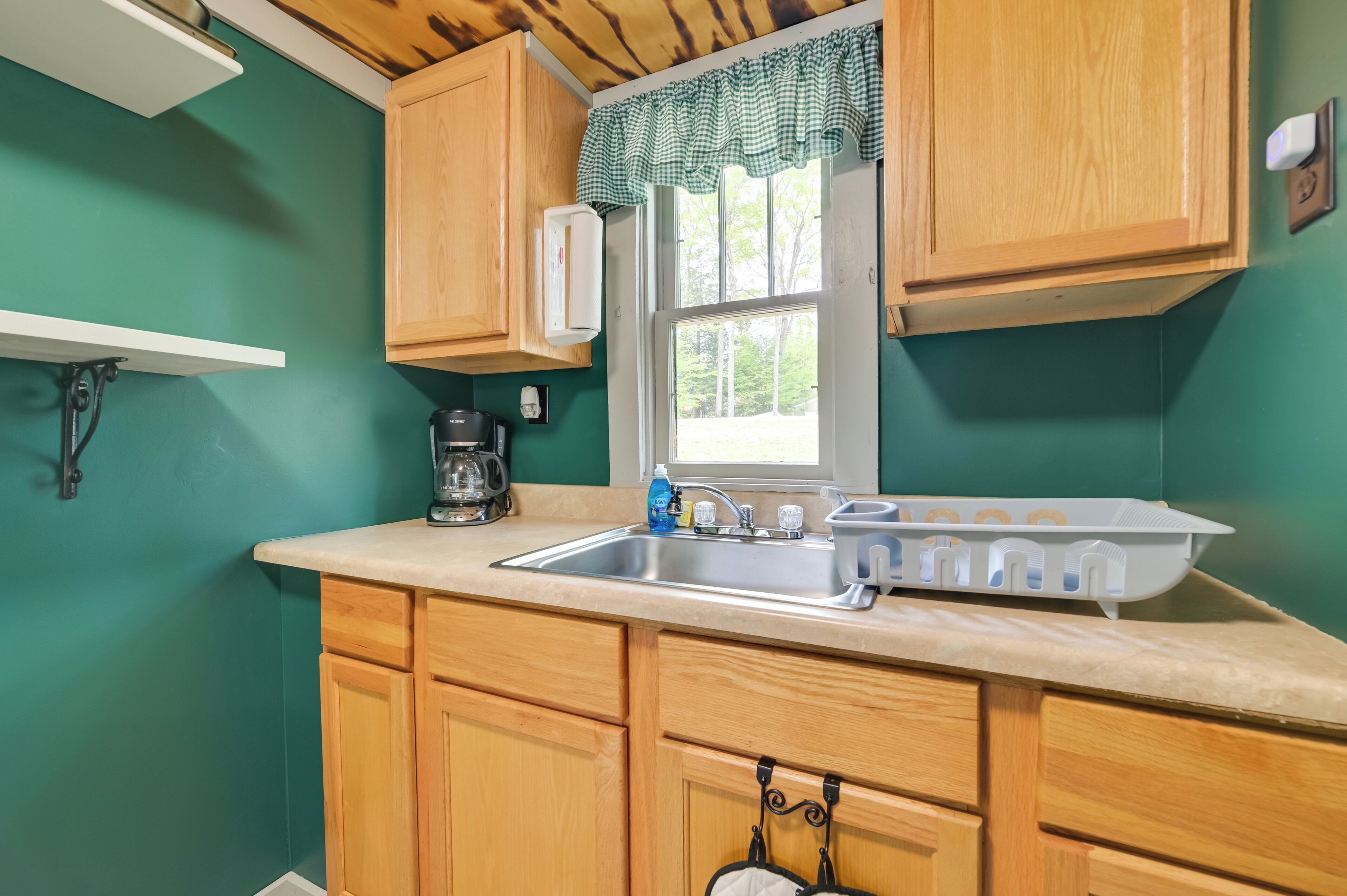 Cocina con armarios de madera, un lavamanos de acero inoxidable, una ventana con cortina a cuadros y una cafetera.