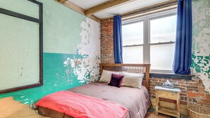 A room with a bed, a wooden headboard, a small table, a wicker basket, and a window with blue curtains. - Direct Trail Access! ATV Riding Home Base in Gary (Gary)