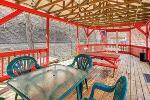 A covered outdoor deck with a table and chairs, wooden railings, and a view of a wooded area.