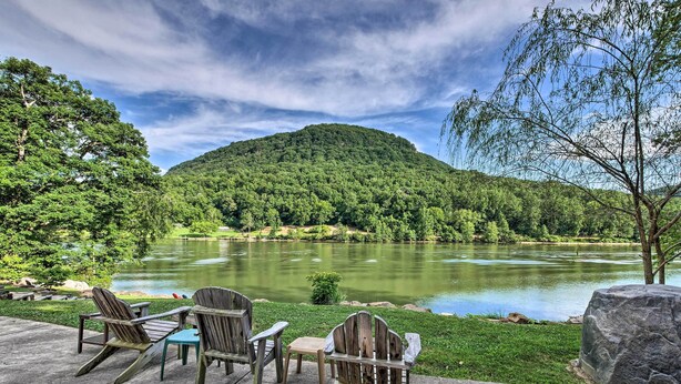 A lakeside seating area with Adirondack chairs and a small table, overlooking a calm lake and a forested hill.