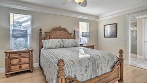 Une chambre avec un lit en bois, deux tables de chevet, un ventilateur de plafond et de grandes fenêtres.