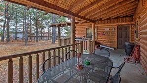 Una cabaña de madera con un porche cubierto, una mesa de comedor con sillas y vistas al bosque circundante.