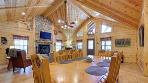 Una cabaña de madera espaciosa con una gran mesa de comedor, varias ventanas, una chimenea de piedra y trofeos de animales colgados.