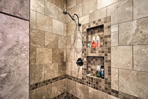 Un baño con azulejos color crema, un cabezal de ducha y un estante con artículos de aseo.