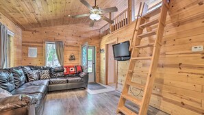 A wooden cabin interior with a leather sofa, a ceiling fan, a TV, and a ladder. - Charming Houghton Lake Cottage w/ Backyard! (Prudenville)