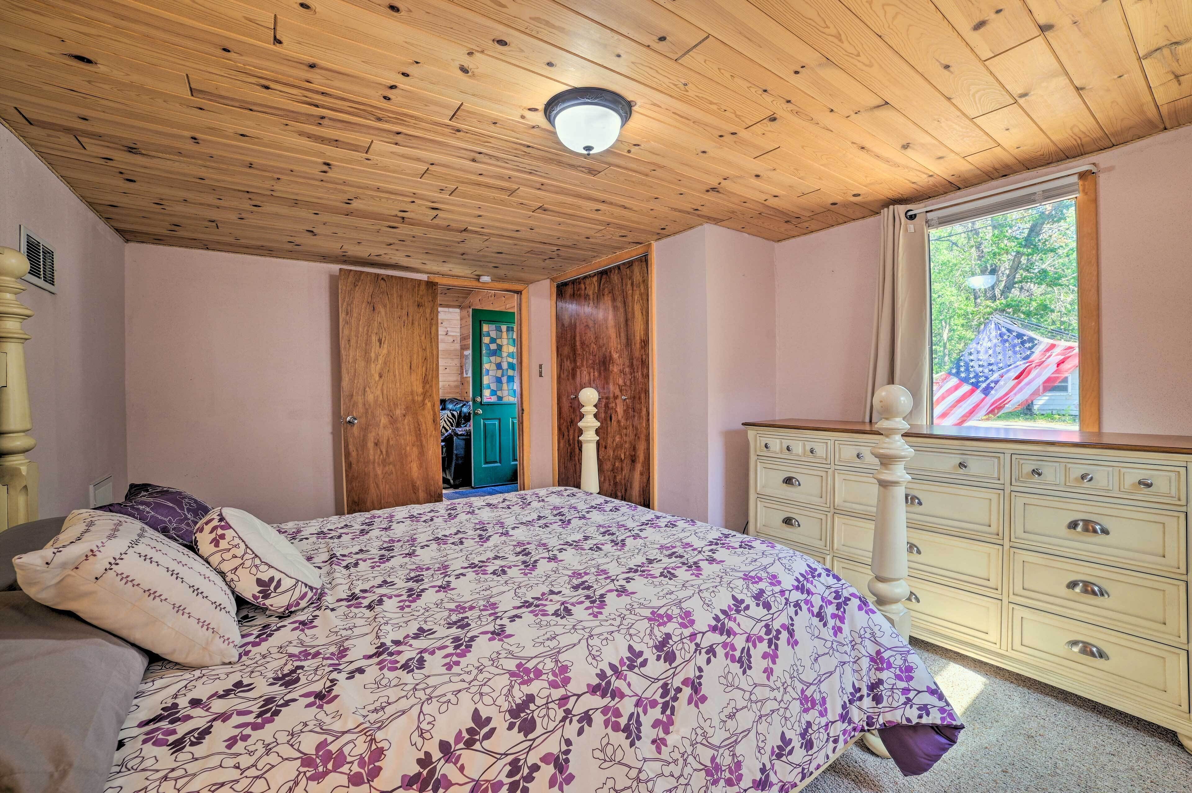 Une chambre avec un plafond en bois, un lit recouvert d’une couvre-lit à motifs floraux, une commode et une fenêtre ornée d’un drapeau américain.