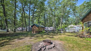 A rustic cabin with a fire pit in the center, surrounded by a wooded area. - Charming Houghton Lake Cottage w/ Backyard! (Prudenville)